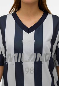 Marineblå og hvitstripet trøye med V-hals, med et sølvfarget logoavtrykk «MILANO» og «98», laget av et teksturert, pustende stoff.