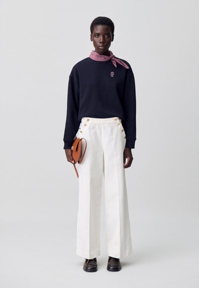 Sudadera negra con logo, pantalones anchos blancos con botones dorados laterales y un bolso cruzado marrón. Bufanda de patrón rojo y blanco atada al cuello.