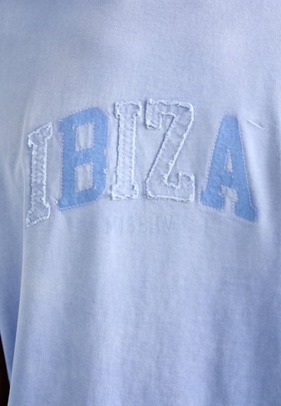 Heleheles sinine puuvillane T-särk, mille ette on kuduma "IBIZA" tekstuurses valges tikandis. Pehme tekstuur ja sile viimistlus, minimalistlik disain.