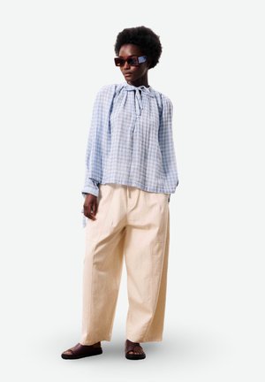 Persoon met donker krullend haar die een oversized lichtblauwe geruite blouse, brede beige broek, donkere sandalen en grote rechthoekige zonnebril draagt, staand.