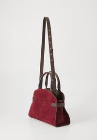 Red suede handbag with brown leather accents, featuring dual handles and a long adjustable strap. Rounded shape and minimalist design.