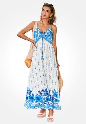 Kobieta w białej i niebieskiej kwiecistej sukience maxi bez rękawów, trzymająca siatkową torbę z pomarańczami i słomkowy kapelusz, stojąca z ręką na biodrze.