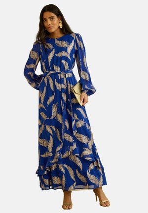 Femeie purtând o rochie lungă albastră cu modele aurii de pene, mâneci bufante, ținând o punguță aurie și purtând tocuri și cercei aurii.