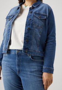 Jeansjacka i medelblå färg med knäppning fram, två bröstfickor och krage, kombinerad med en vit skjorta och blå jeans.