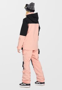 Roze en zwarte sneeuwjack met capuchon, gecombineerd met bijpassende roze sneeuwbroek. Voorzien van zijzakken en zwarte accenten. Zwart-witte beanie op het hoofd.
