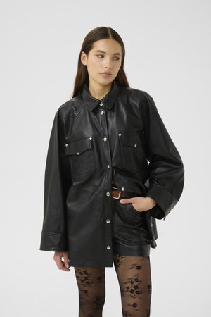 Giacca camicia oversize in pelle nera con bottoni a pressione, tasche sul petto e colletto appuntito; indossata sopra collant neri a fantasia.