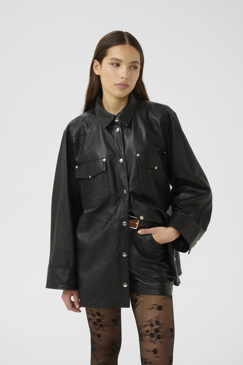Giacca camicia oversize in pelle nera con bottoni a pressione, tasche sul petto e colletto appuntito; indossata sopra collant neri a fantasia.