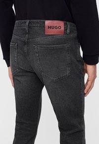 Homme portant un jean noir avec un patch logo HUGO rouge sur la ceinture arrière, vu de dos de la taille jusqu'à mi-cuisse.