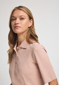 Koszulka polo w jasnoróżowym kolorze, wykonana z teksturowanej tkaniny. Posiada kołnierzyk i krótkie rękawy, z subtelnym logo wyhaftowanym na rękawie.