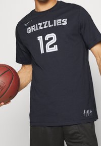 Tmavě modrý sportovní dres s nápisem "GRIZZLIES" a číslem "12" v bílé barvě. Obsahuje síťovou texturu a logo Nike. Podpis hráče na lemu.