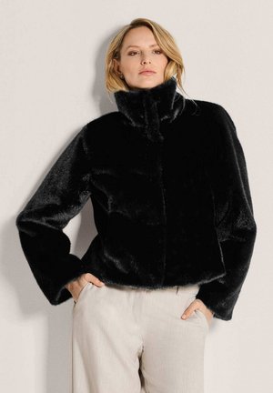 Frau trägt eine schwarze Jacke aus Kunstfell mit hohem Kragen und beige Hose mit Händen in den Taschen vor einfarbigem Hintergrund.