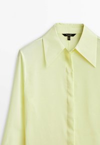 Camicia a maniche lunghe gialla chiara con colletto strutturato, chiusura con bottoni e texture liscia; design minimalista senza motivi o dettagli.