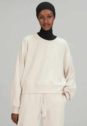 Helles beiges Sweatshirt mit lockerem Schnitt, Rundhalsausschnitt, überschnittenen Schultern und gerippten Bündchen. Kombiniert mit passenden, schmal zulaufenden Hosen.