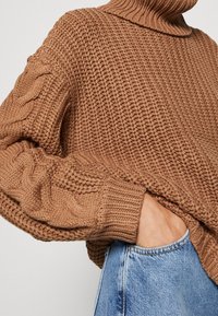 Maglione marrone a maglia con colletto alto, caratterizzato da dettagli a treccia sulle maniche. Indossato sopra jeans in denim blu chiaro.