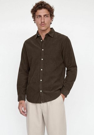 Jeune homme aux cheveux bruns bouclés portant une chemise boutonnée marron foncé et un pantalon beige clair, debout avec une main dans la poche.