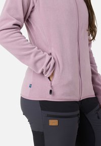 Fleece-takki pehmeässä vaaleanpunaisessa, jossa on täysi vetoketju, sivutaskut ja pieni sininen ja keltainen etiketti. Yhdistetty tummiin, tekstuurisiin housuihin.