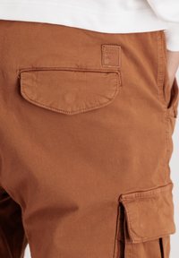 Pantalons cargo marron en coton avec une poche arrière. Présente une étiquette en cuir rectangulaire avec un texte en relief. Texture lisse.