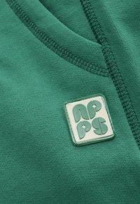 Close-up van groene stof met gedetailleerde stiksels en een vierkante rubberen patch met abstracte vormen in bijpassend groen.