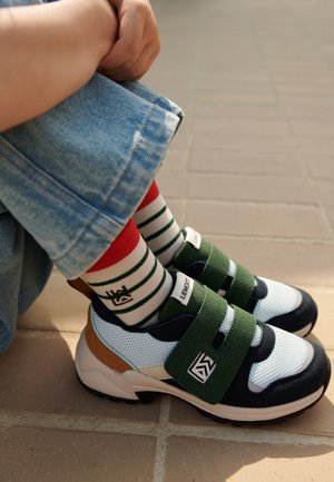 Sneaker multicolori con mesh blu, cinture verde e nera e un accento di tacco marrone; abbinate a calzini a righe rosse e bianche.