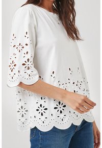 Blouse blanche avec des motifs floraux découpés complexes le long de l'ourlet festonné et des manches, associée à une coupe décontractée et une texture douce.