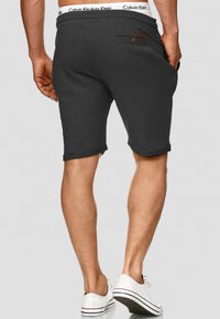 Grau Shorts aus weichem Stoff, mit umgeschlagenen Säumen, einer Gesäßtasche mit Knopf und einem elastischen Bund mit Logo. Dazu werden weiße Schuhe getragen.