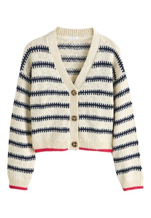 Mosolygó gyerek hosszú szőke hajjal csíkos kardigánt, piros inget, bő farmernadrágot és fekete tornacipőt visel, és egy világos ablak mellett áll.