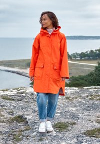 Long orange raincoat with a hood, featuring front pockets and a slightly curved hem. Paired with light blue rolled-up jeans and white sneakers.