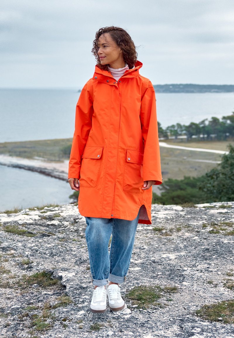 Langer orangefarbener Regenmantel mit Kapuze, vorne mit Taschen und einem leicht abgerundeten Saum. Kombiniert mit hellblauen, hochgekrempelten Jeans und weißen Turnschuhen.