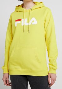 Žlutá fleecová mikina s kapucí, přední kapsou, šňůrkami na kapuci a bílým a červeným logem FILA přes hruď.