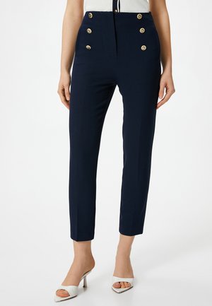 Pantaloni a vita alta blu navy con dettagli in bottone dorato, gamba affusolata e un tessuto liscio e testurizzato. Tacchi bianchi corti visibili.