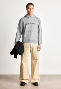 Muž v šedé mikině Calvin Klein, béžových širokých kalhotách, držící černou bundu, stojící na šedé podlaze proti jednoduché bílé zdi.