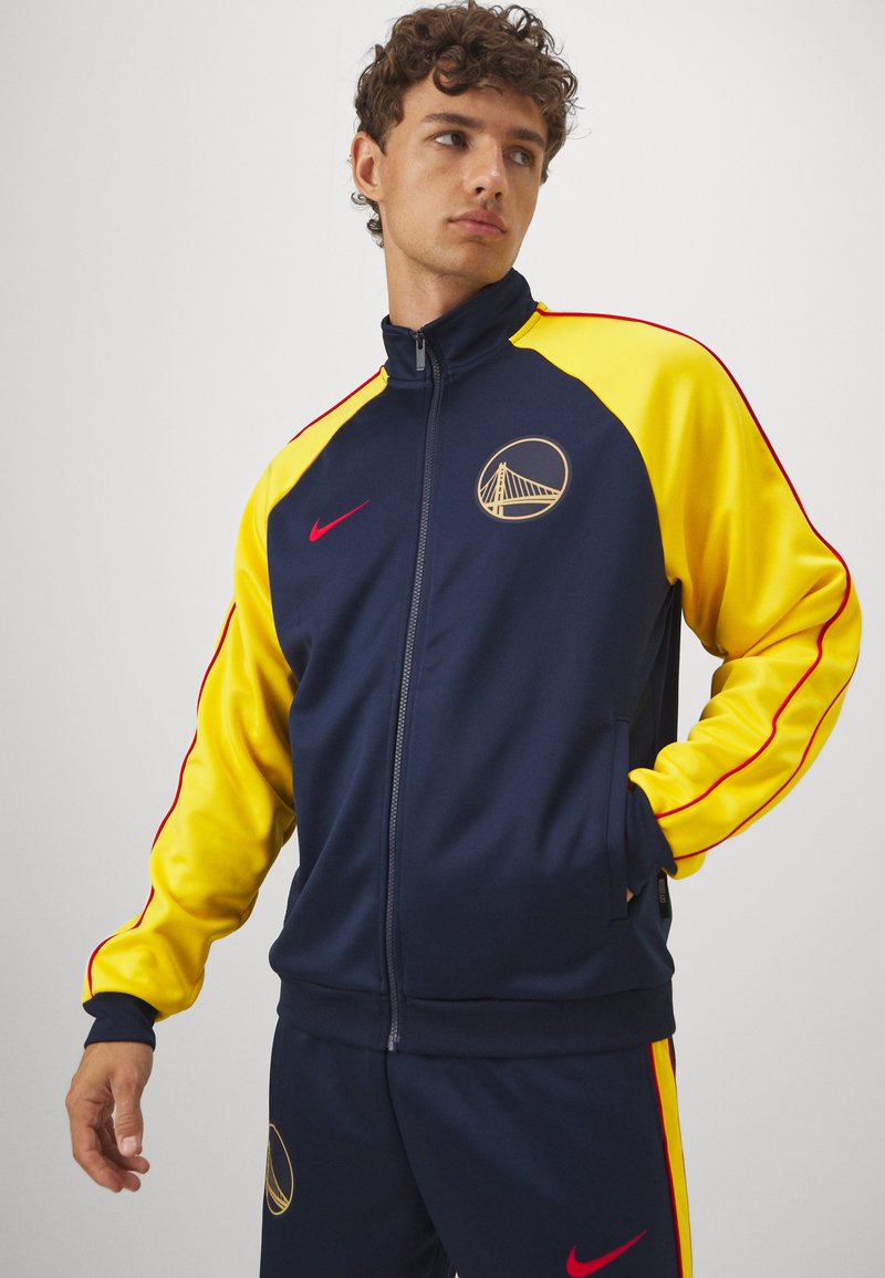 Giacca da tuta blu navy e gialla con zip completa, dettagli rossi e logo dei Golden State Warriors. Realizzata in un materiale sportivo e morbido.
