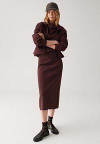 Bordeauxfarbener Pullover mit eng anliegenden Ärmeln, passendem Midirock, kombiniert mit schwarzen Loafers und transparenten schwarzen Socken, abgerundet mit einer grauen Mütze.