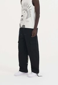 Schwarze Cargo-Hosen mit Seitentaschen, mit buntem gesticktem Design. Kombiniert mit einem ärmellosen weißen Tanktop mit Grafikdruck. Weiße Sneaker.