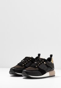 Czarno-złote sneakersy z siateczkową cholewką, zamszowymi akcentami, złotymi detalami oraz masywną białą i jasnobrązową podeszwą.