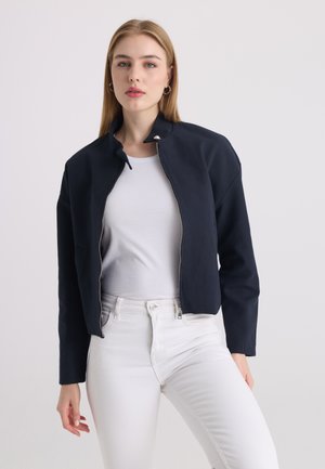 Femme portant une veste zippée bleu marine par-dessus une chemise blanche et un pantalon blanc, debout devant un fond clair uni.