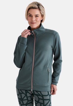 Frau trägt eine türkisfarbene Jacke mit Reißverschluss und gemusterte Leggings, hält den Reißverschlussgriff in der Nähe des Halses, schaut nach vorne und lächelt leicht.