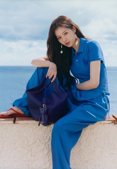 Mladá žena v modrém polu a kalhotách sedí na zdi u moře a drží velkou tmavě modrou kabelku s medvídkovým přívěskem na klíče.