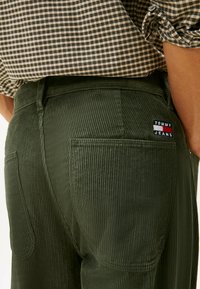 Olīvu zaļi korduroja bikses ar vertikālu ribotu tekstūru, ar aizmugurējo kabatu un zīmolu plāksni ar "TOMMY JEANS" sarkanā un zili baltā krāsā.