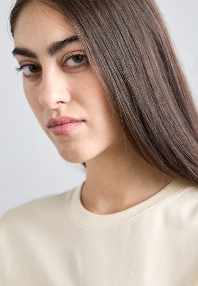 Beige sweatshirt with a round neckline, soft texture, and visible stitching. Long, dark hair partially falling over shoulders. Natural makeup.