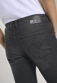 Närbild av en person som bär svarta Tom Tailor denimjeans med bakfickor och varumärkesmärke, tillsammans med en vit skjorta.