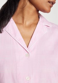 Różowa koszula w kratę z kołnierzykiem, delikatną strukturą, zapięciem na guziki i białym wykończeniem na krawędziach.