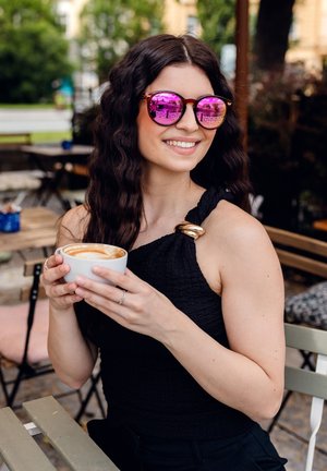 Černý texturovaný top s akcentem na rameni, růžové zrcadlové sluneční brýle, bílá miska s kávou, ruce na zeleném stole v prostředí venkovní kavárny.