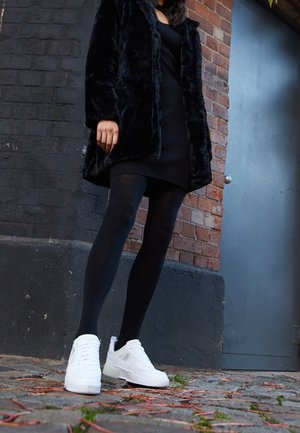 Woman wearing black dress, black tights, black fur coat, and white sneakers standing on cobblestone pavement near brick wall and blue door.