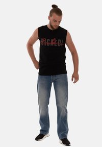 Schwarzes ärmelloses T-Shirt mit rotem und grauem grafischem Text. Kombiniert mit hellblauen Jeans und schwarzen Turnschuhen mit weißen Sohlen. Lässiger Schnitt.