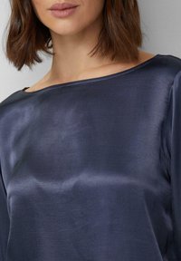 Marineblauwe satijnen top met een brede halslijn en een gladde textuur. Heeft een ontspannen pasvorm en minimale stikwerkdetails langs de randen.