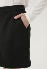 Svart mockakjol med en slät yta, rak passform, sidofickor och midjeband. Matchas med en långärmad vit topp.