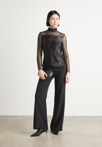 Schwarzes Paillettentop mit durchsichtigen langen Ärmeln, kombiniert mit hoch sitzenden schwarzen Hosen und schwarzen spitzen Stiefeletten. Enthält eine kleine schwarze Clutch.