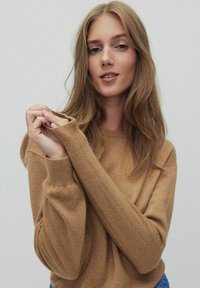 Pull en couleur camel en maille douce, avec des manches longues et des poignets côtelés, porté de manière décontractée avec une coupe ample.