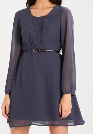 Vrouw draagt een marineblauwe jurk tot op de knie met doorschijnende lange mouwen, plooien aan de voorkant en een dunne zwarte riem in de taille.